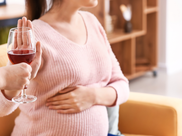 Een zwangere vrouw met een roze shirt aan zegt nee tegen een glas rode wijn. Ze zegt nee omdat ze zwanger is en zwangere vrouwen mogen geen alcohol drinken.