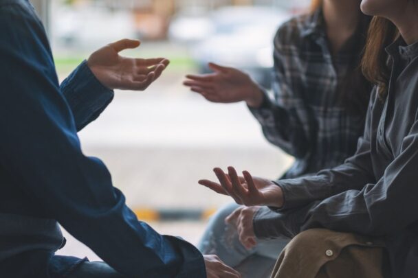 Drie personen zitten bij elkaar. Ze bewegen met hun handen naar elkaar