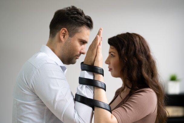 Een man en vrouw zijn vastgebonden aan hun polsen. Ze kijken elkaar aan.