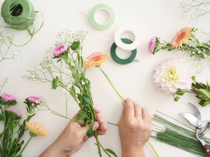 Twee handen pakken losse bloemen bij elkaar. Op tafel liggen meer bloemen en lint.