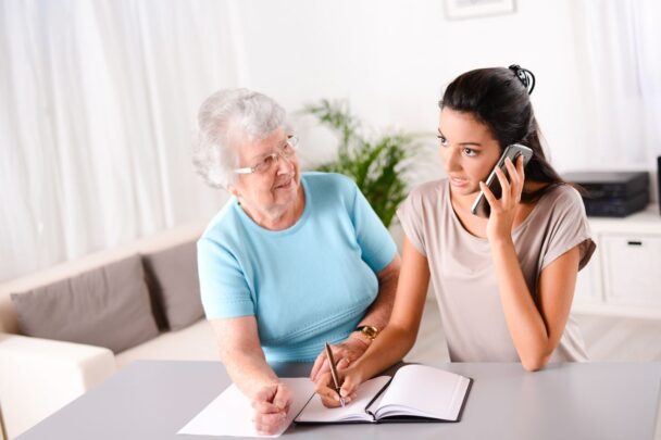 Jongere vrouw is aan het bellen en in een schrift aan het schrijven. Een oudere vrouw kijkt naar haar.