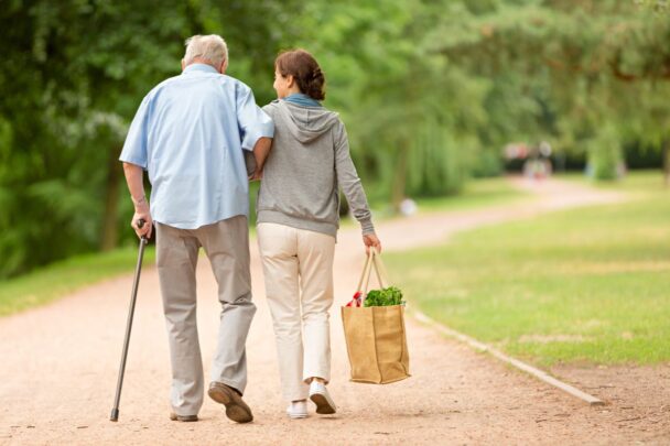 Man en vrouw wandelen buiten. Man heeft wandelstok vast, vrouw draagt boodschappentas en ondersteunt man.