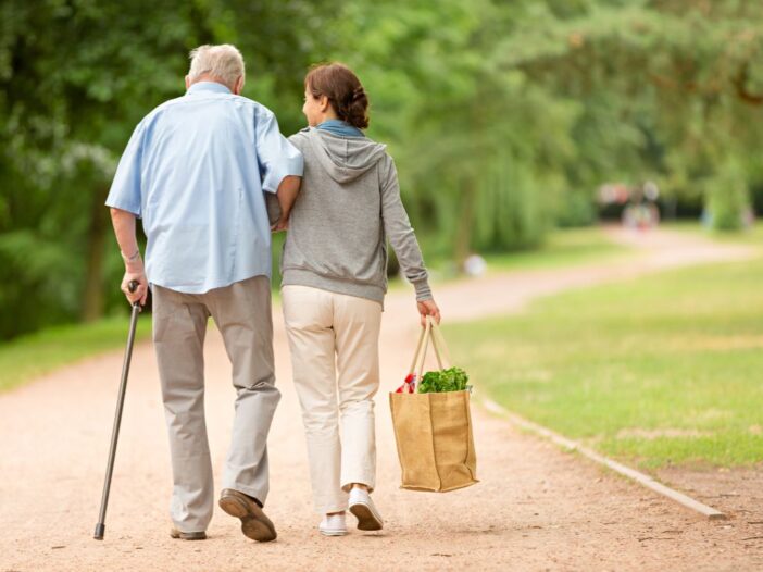 Man en vrouw wandelen buiten. Man heeft wandelstok vast, vrouw draagt boodschappentas en ondersteunt man.