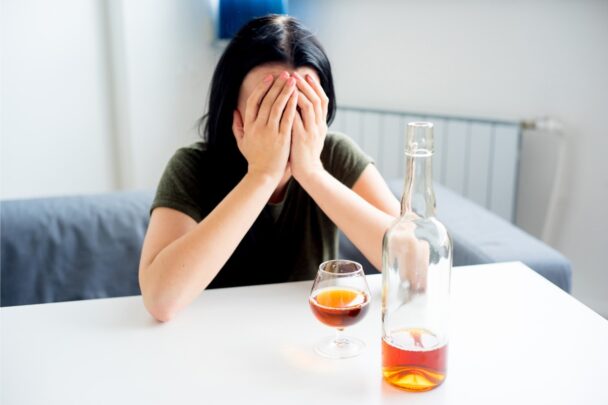 Vrouw met haar handen voor haar gezicht. Op tafel staat een fles drank en een volgeschonken glas.