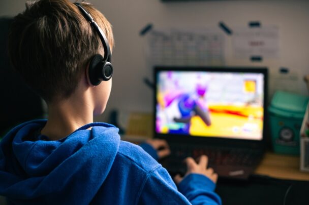 Achteraanziht van een kind met koptelefoon op. Hij speelt een videogame op een laptop.