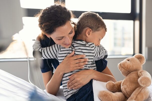 Vrouw geeft een jong kind een knuffel. Hij zit op een ziekenhuisbed met een teddybeer naast hem.