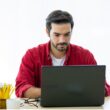 Man met rode blouse aan maakt gebruik van een laptop. Hij zit aan een tafel.