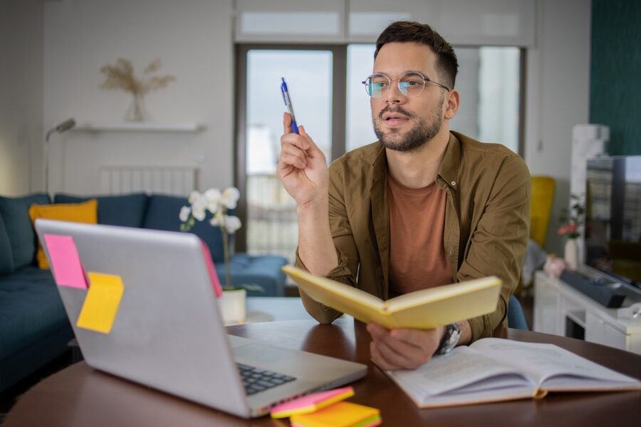 Man met bril zit achter laptop met een boek en pen in zijn handen.