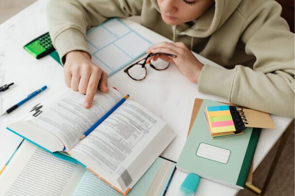 Meisje buigt zich over een tafel met een boek, bril en wat schoolspullen erop