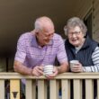 Oudere man en vrouw staan samen op een veranda en genieten van een kop koffie.