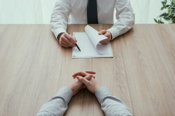 Twee mannen in pak zitten aan tafel in een serieus gesprek. De een houdt papieren vast en een pen.