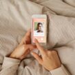 Een paar handen houdt een smartphone vast waarop datingapp te zien is. De smartphone ligt op een dekbed.