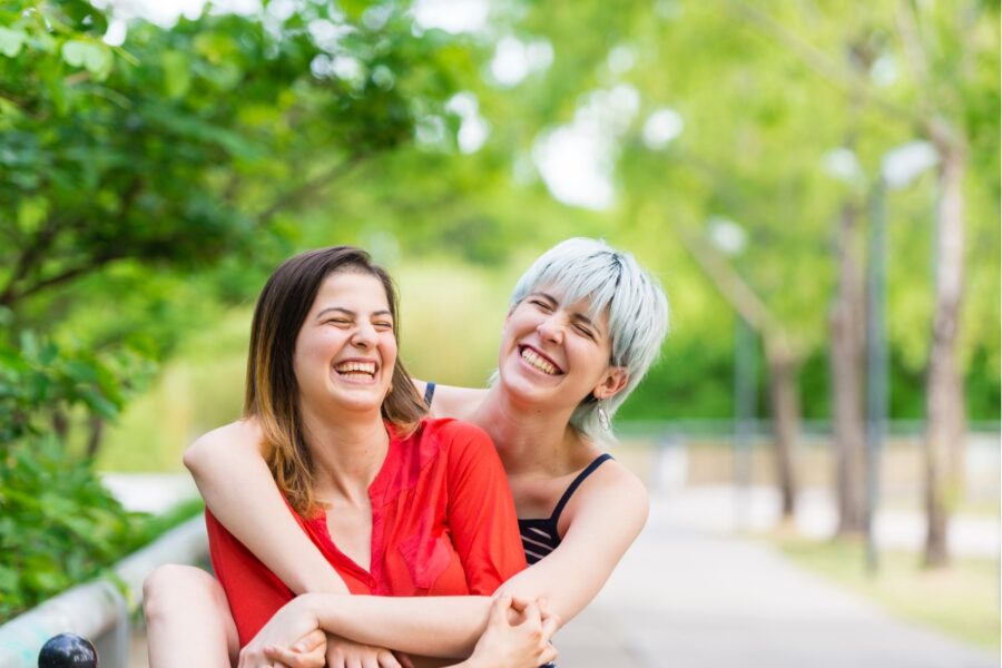 Twee verliefde jonge vrouwen die elkaar vasthouden. Ze kijken blij. De achtergrond is groen en rijk aan bomen.