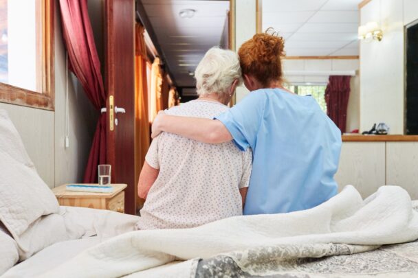 Achteraanzicht van twee vrouwen op bed. De een heeft een arm om de ander heen geslagen.