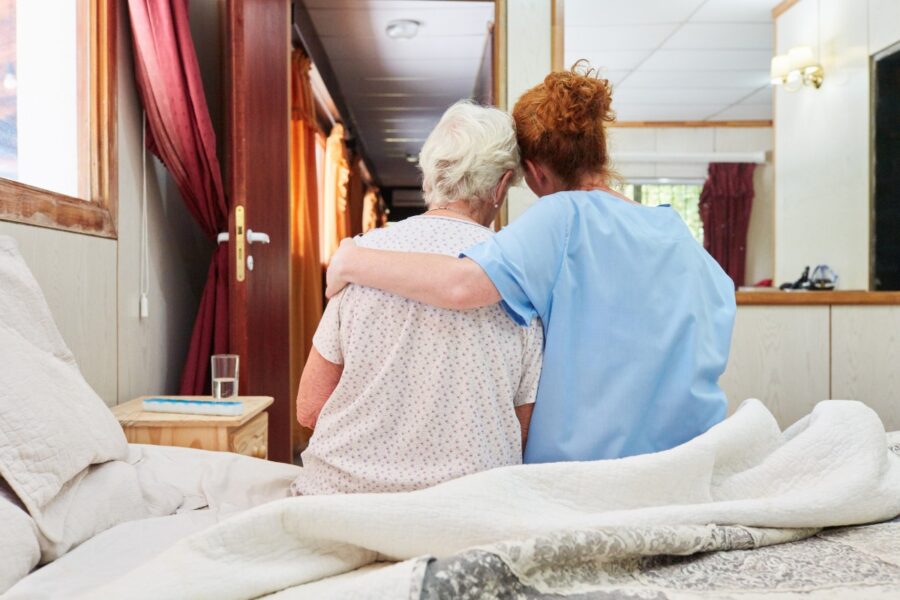 Achteraanzicht van twee vrouwen op bed. De een heeft een arm om de ander heen geslagen.