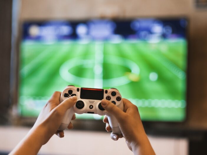 Twee handen houden een game controller vast. Op het beeldscherm zie je een voetbalspel.