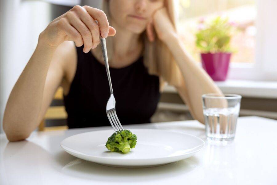 Jonge vrouw zit aan een eettafel met voor haar een bord. Er ligt 1 roosje broccoli op het bord.