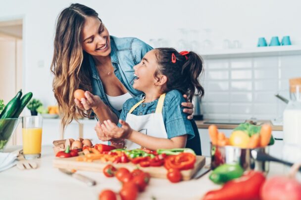 Een jonge vrouw en een meisje bereiden samen een gezonde maaltijd in de keuken. Ze kijken elkaar lachend aan.