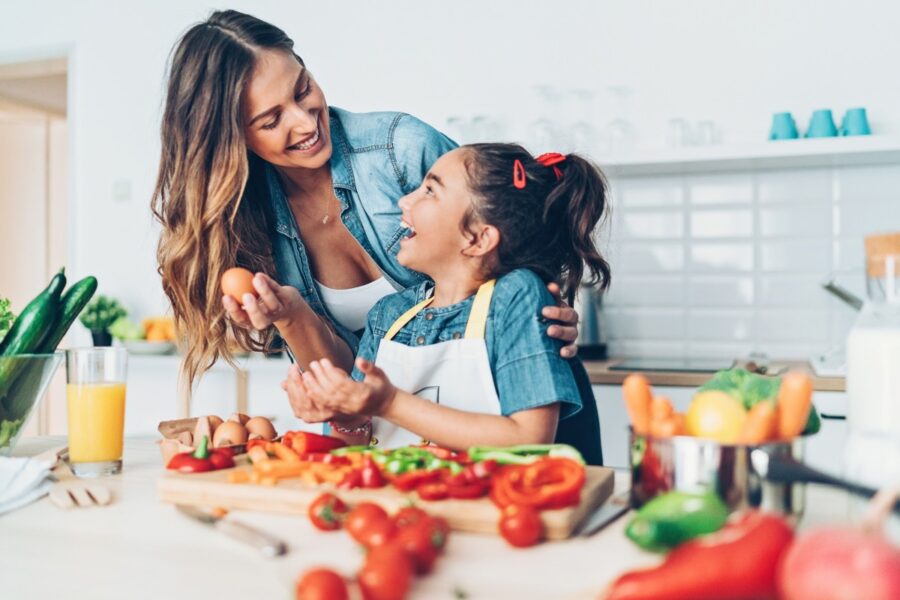 Een jonge vrouw en een meisje bereiden samen een gezonde maaltijd in de keuken. Ze kijken elkaar lachend aan.