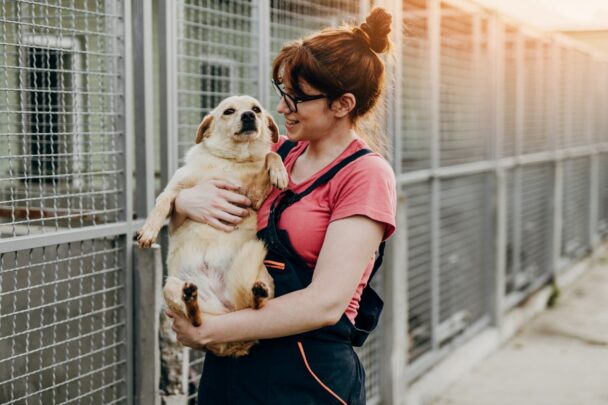 Vrouw houdt een puppy in haar armen bij een asiel. Op de achtergrond zie je de kooien van andere dieren.