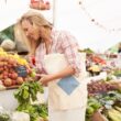 Blonde, jonge vrouw loopt met een canvas tas op een markt. Ze bekijkt groenten en fruit.