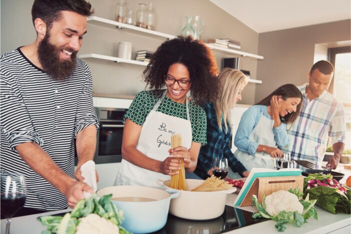 Drie vrouwen en twee mannen koken samen een maaltijd. De sfeer is gezellig.