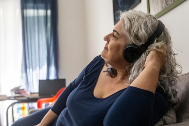Oudere vrouw luistert op de bank naar iets via een koptelefoon. Ze oogt ontspannen.