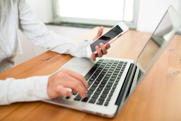 Iemand zit aan een tafel en maakt gebruik van laptop en een smartphone.
