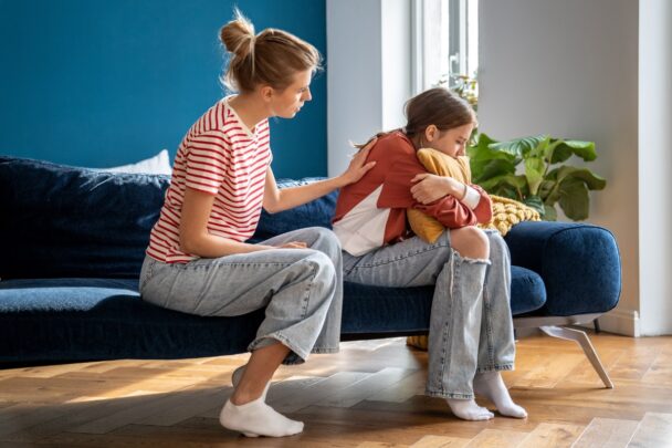 Moeder zit met dochter op de bank. Dochter oogt verdrietig of boos. Ze zit in elkaar gedoken