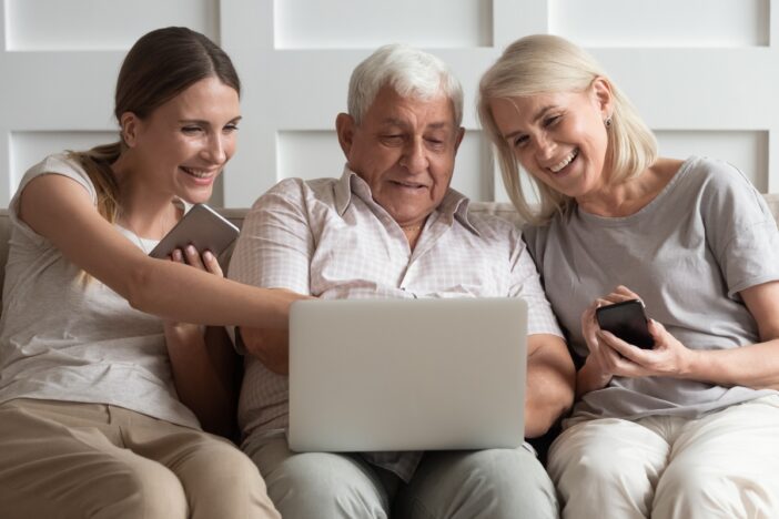 Oudere man en vrouw maken samen met een jonge vrouw gebruik van een laptop, vanaf een bank in een huiskamer.