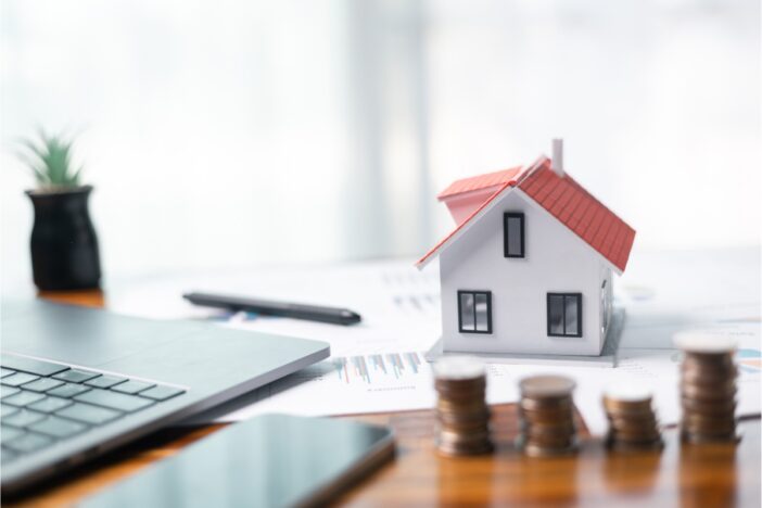 Stapeltje muntgeld op een tafel met daarop een klein papieren huisje en een laptop.