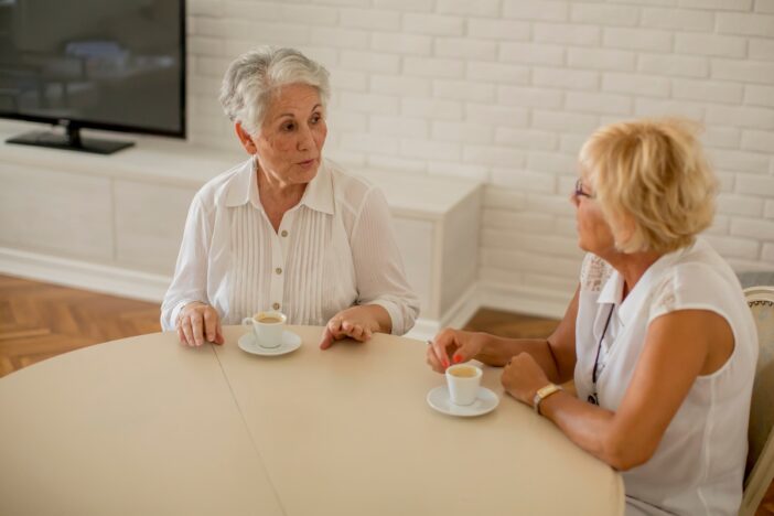 Twee oudere dames drinken koffie aan een tafel en voeren een gesprek.