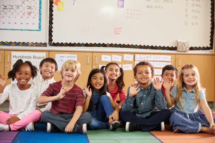 Groepje basisschoolkinderen zit op de grond op een rij en zwaait naar de camera.