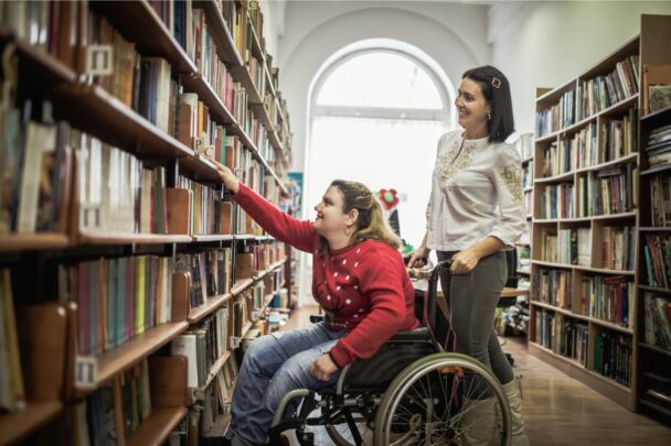 Vrouw in rolstoel pakt een boek uit een kast in een bibliotheek.