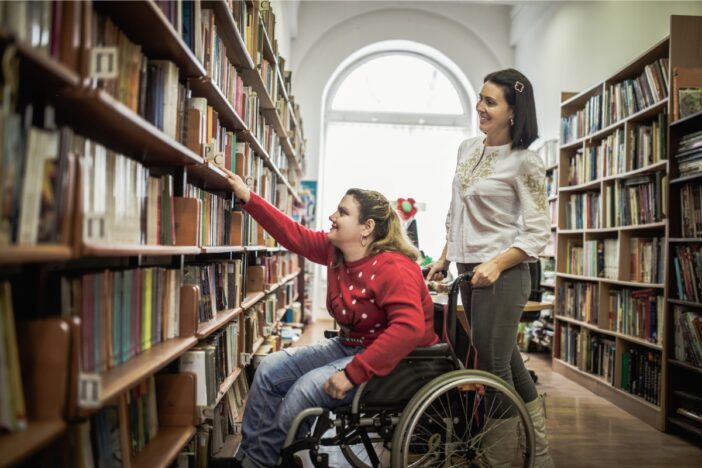 Vrouw in rolstoel pakt een boek uit een kast in een bibliotheek.