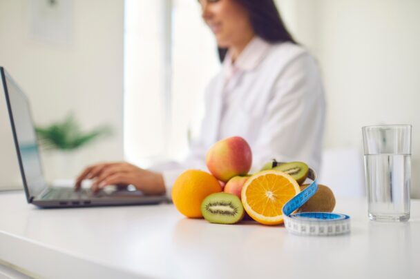 Fruit en een meetlint op een aanrecht. Op de achtergrond zie je een vrouw die een laptop gebruikt.