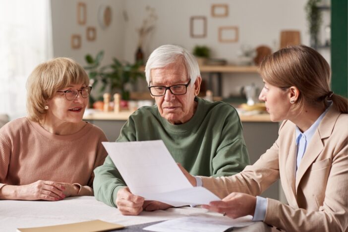 Oudere man en vrouw zitten met een vrouw aan tafel en bespreken een papieren document.