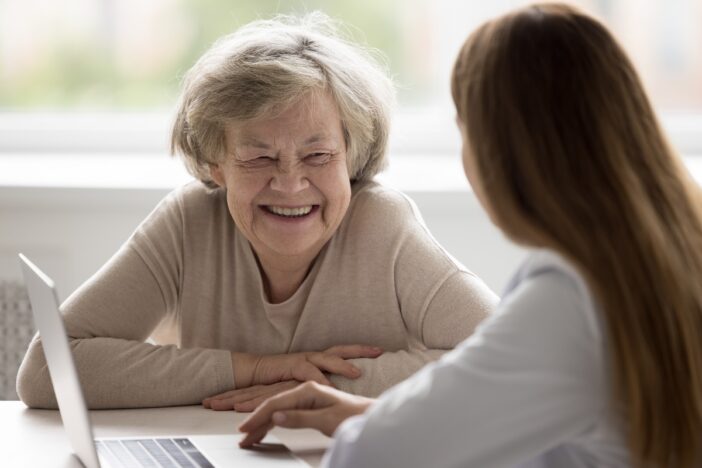 Oudere dame lacht breeduit. Ze zit aan tafel met een jonge vrouw.